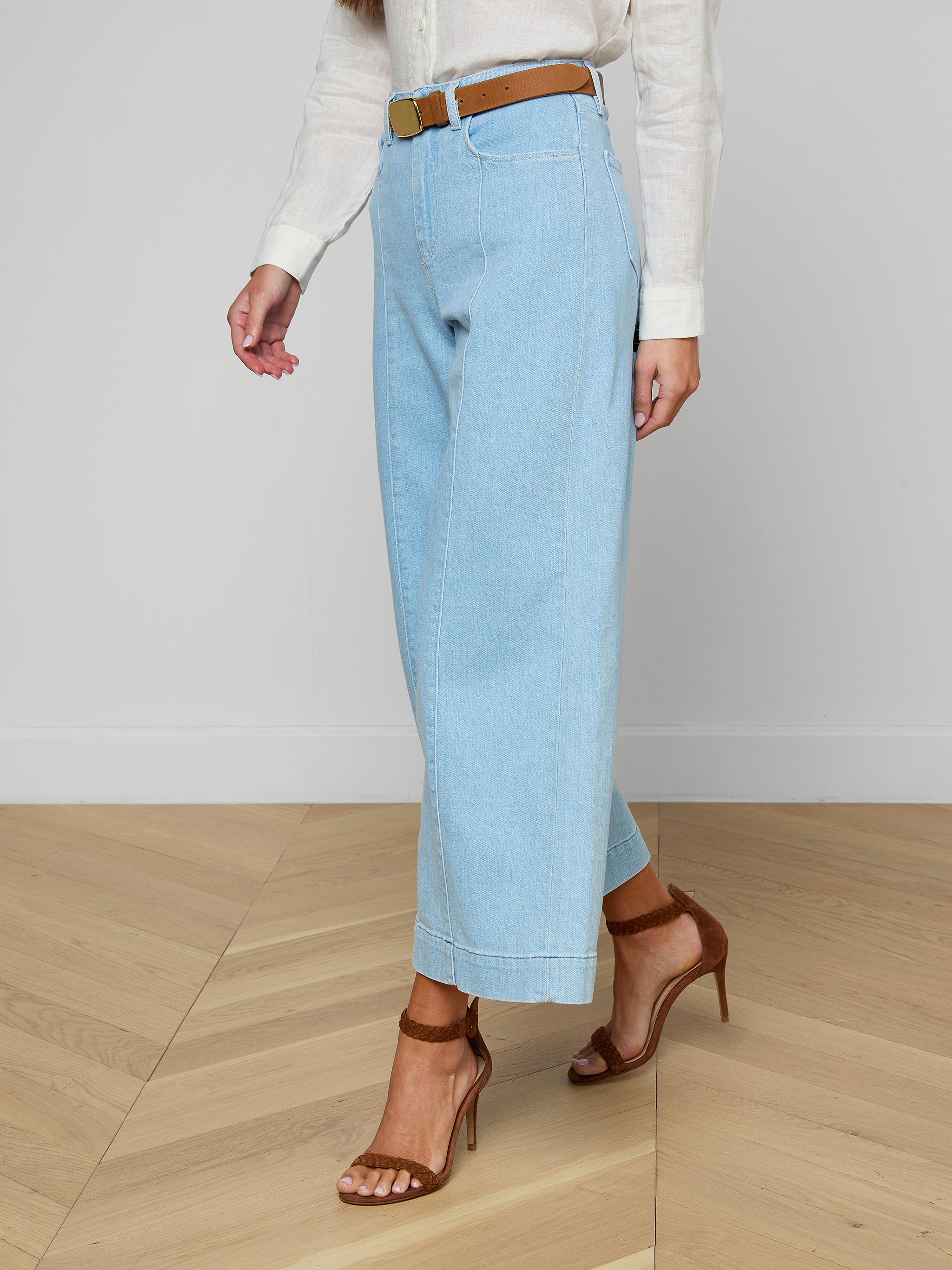 A person is wearing the Houston Cropped Wide-Leg Jeans in light blue stretch denim, paired with a white long-sleeve shirt, a brown belt, and brown high-heeled sandals. Only the lower half of their body is visible against a plain light background.