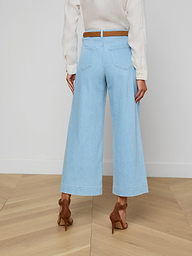 A woman is shown from the back against a plain background, wearing the Houston Cropped Wide-Leg Jean in light blue stretch denim, styled with a white long-sleeve top, a brown belt, and brown high-heeled shoes.