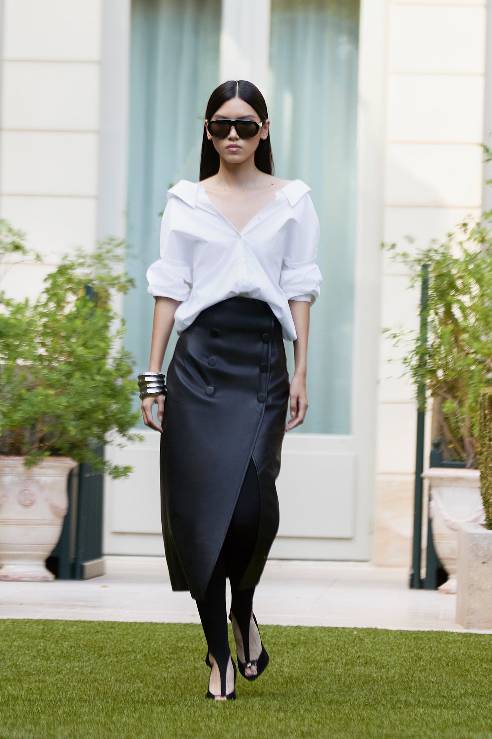 Wearing sunglasses, a white button-up shirt, a long black skirt, and black heels, a person strolls on grass in front of a building with large windows and potted plants.