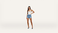 A woman stands against a plain white background, wearing a white tank top, denim shorts, and blue heeled sandals. One hand rests on her hip while the other hangs by her side.