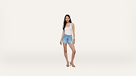 A woman stands against a plain white background, wearing a white tank top, light blue denim shorts, and sandals. She has long dark hair and is looking slightly to the side.
