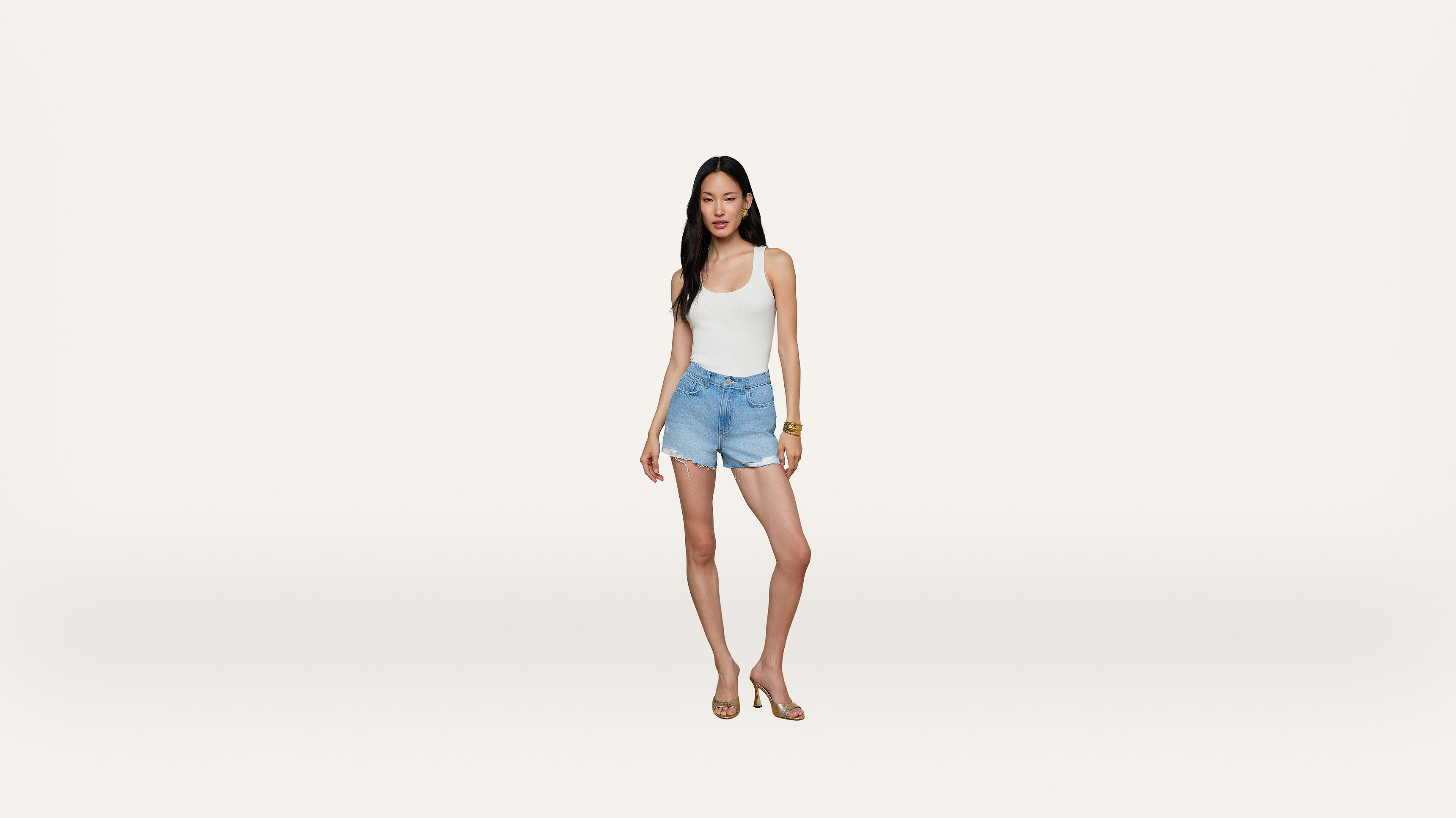 A woman stands against a plain white background, wearing a white tank top, light blue denim shorts, and sandals. She has long dark hair and is looking slightly to the side.