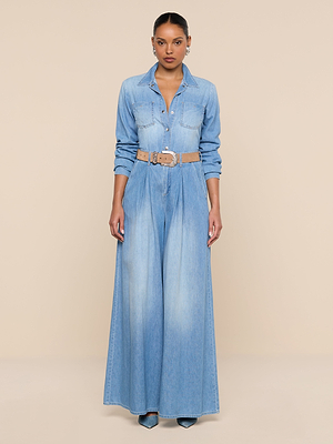 A woman stands facing forward in the Lorenza Palazzo Jean, a featherweight chambray denim jumpsuit with long sleeves, high-rise palazzo legs, and a beige belt. Her hair is pulled back and she wears pointed-toe heels against a plain beige background.