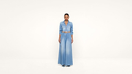 A person stands facing forward, wearing a long denim dress with a belt, against a plain white background.
