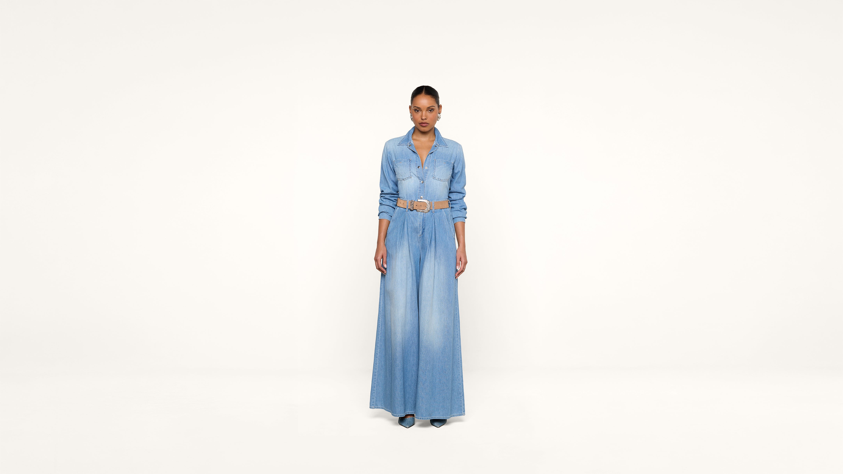 A person stands facing forward, wearing a long denim dress with a belt, against a plain white background.