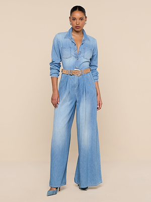 A woman stands against a beige background wearing the Lorenza Palazzo Jean, a high-rise, wide-leg denim pant paired with a light blue chambray jumpsuit top, beige belt, blue heels, hoop earrings, and her hair pulled back.
