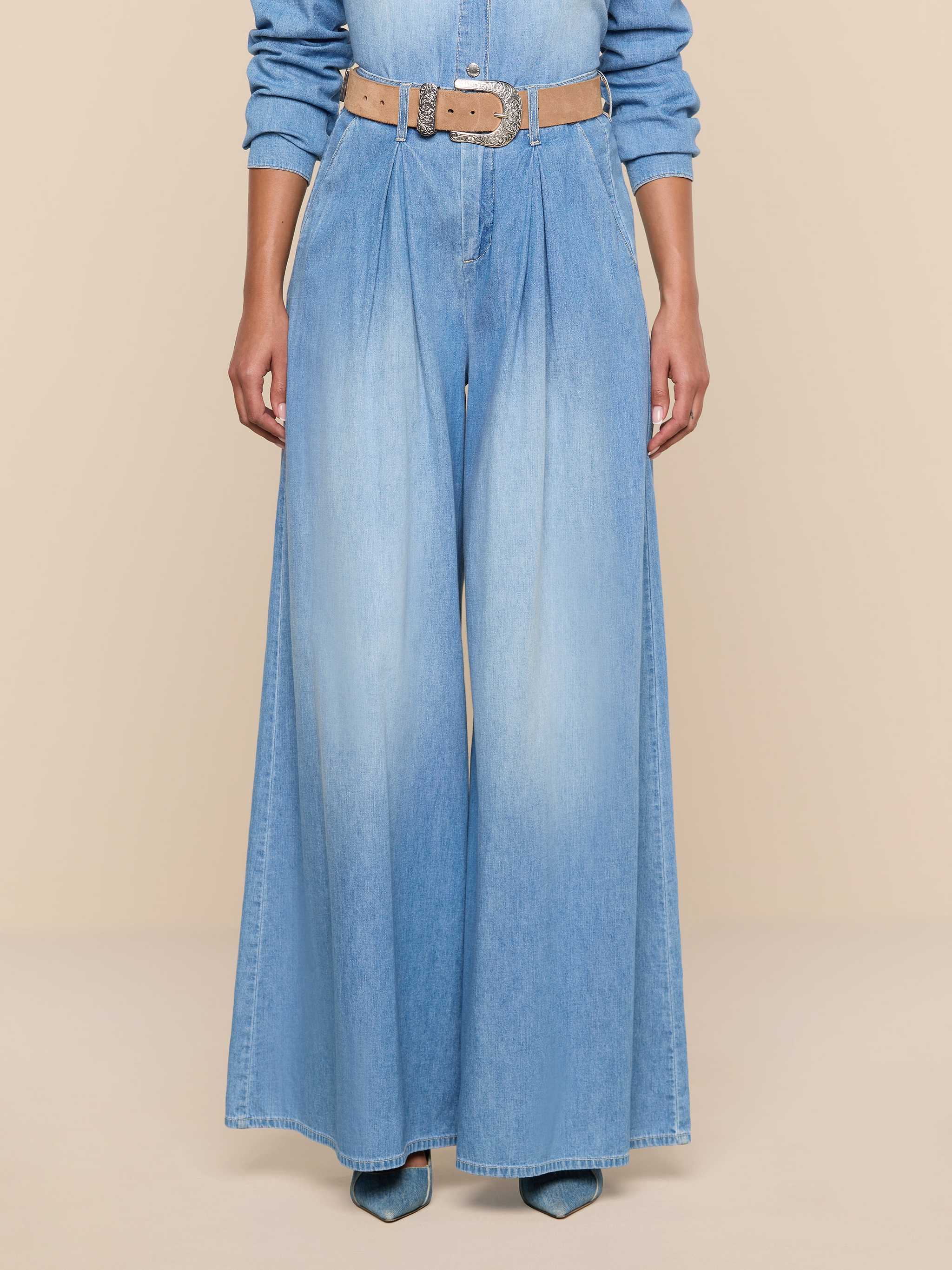Wearing the Lorenza Palazzo Jean in light blue denim, paired with a tan belt and pointed-toe blue shoes, a person is shown from the shoulders down against a plain white background.
