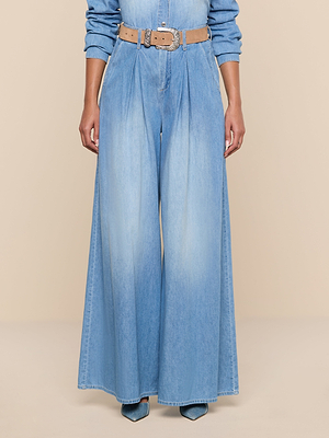 A person wears the Lorenza Palazzo Jean—high-waisted, featherweight chambray wide-leg jeans with pleats—styled with a tan belt and pointed blue shoes, pictured partially against a neutral beige background.