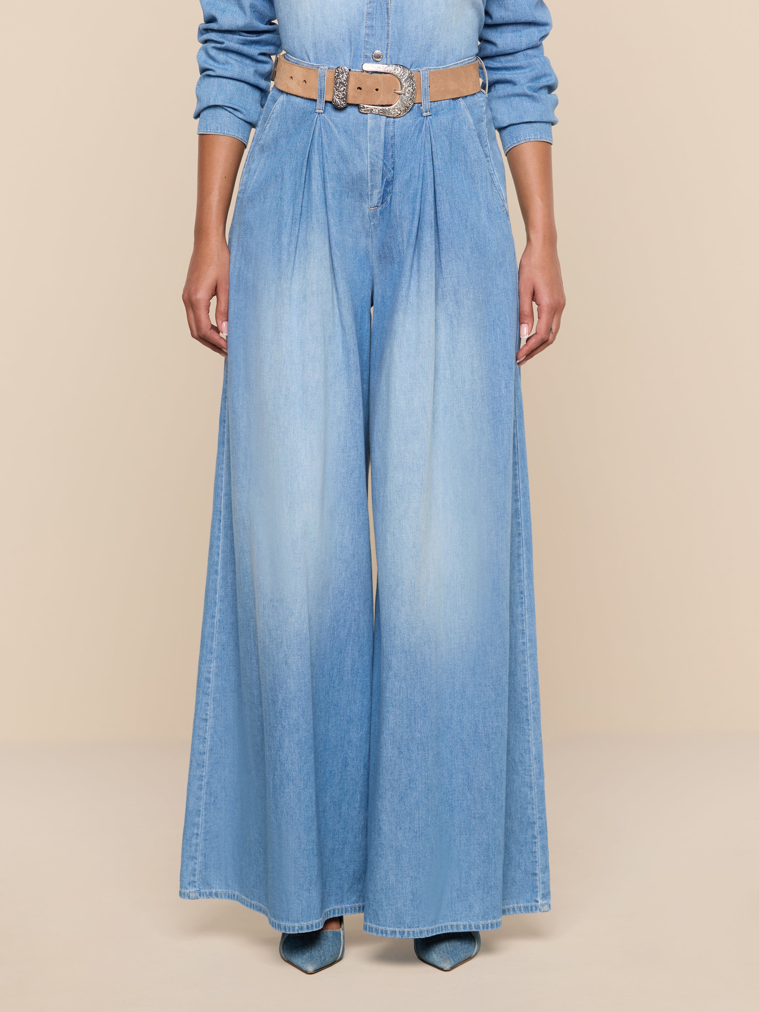 Wearing the Lorenza Palazzo Jean in light blue denim, paired with a tan belt and pointed-toe blue shoes, a person is shown from the shoulders down against a plain white background.