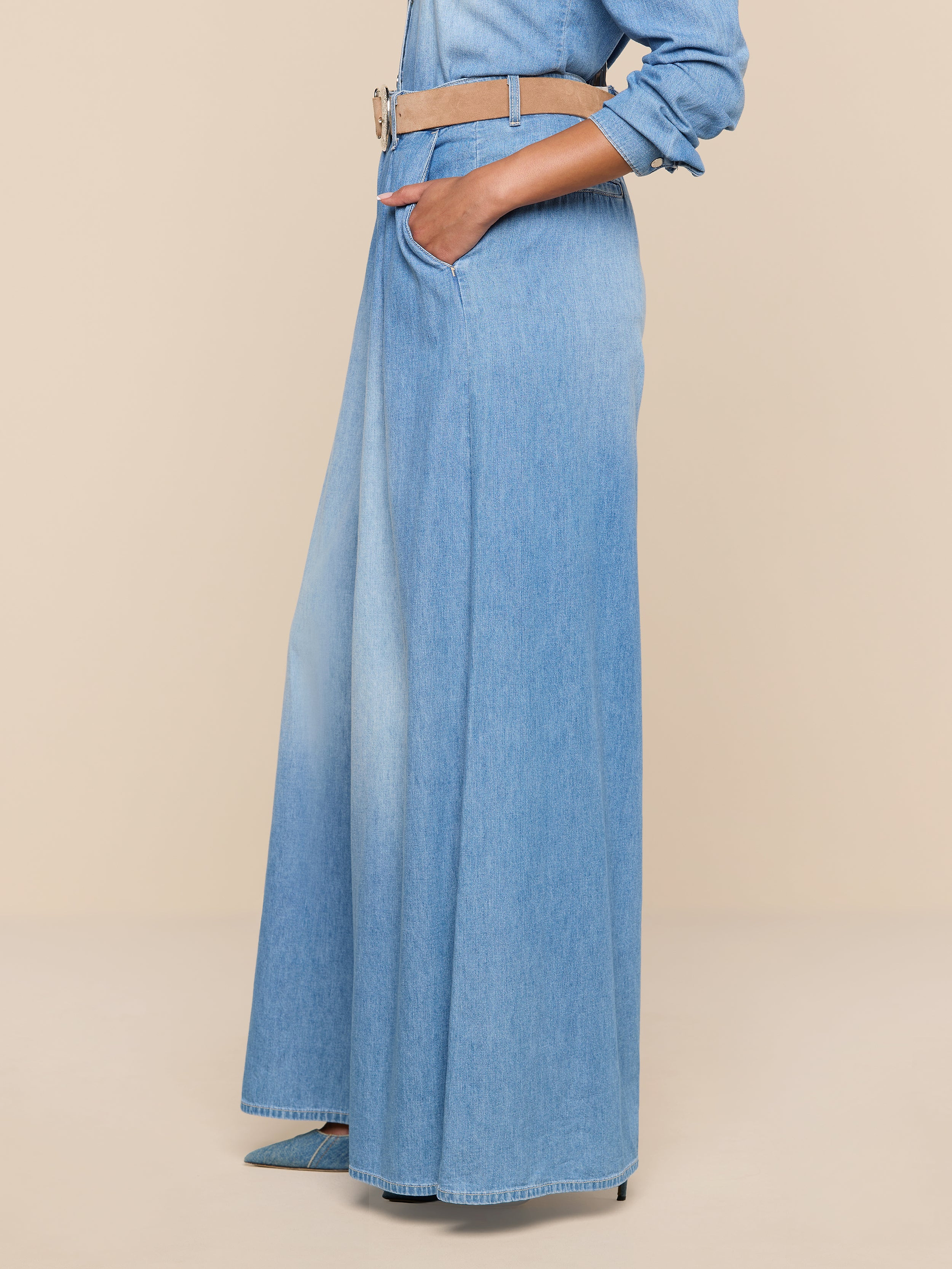 A person is wearing the Lorenza Palazzo Jean in light blue featherweight chambray denim, styled with a wide tan belt, hands in pockets, and a matching denim shirt. The background is plain white.