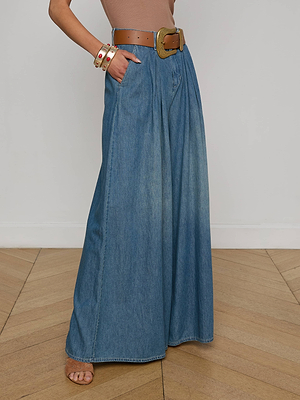 A person stands on a wooden floor against a plain white wall, wearing the Lorenza Palazzo Jean, a tan sleeveless top, a wide brown belt, two bracelets, and open-toed sandals.