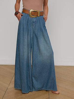 Wearing the Lorenza Palazzo Jean in a vintage wash with a brown belt featuring a large gold buckle, a sleeveless beige top, and bracelets, standing on a wooden floor against a plain white wall.