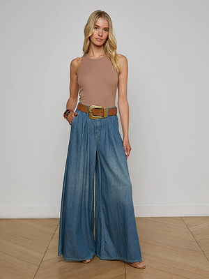 A woman with long, wavy blonde hair stands indoors wearing a sleeveless beige top, Lorenza Palazzo Jean in vintage wash with a wide brown belt, bracelets, and sandals. She poses with one hand in her pocket.
