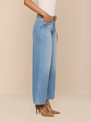 Una persona lleva el Alicent Wide-Leg Jean en un lavado índigo claro, combinado con un top blanco, cinturón beige y tacones marrones de punta. La foto muestra solo la parte inferior del cuerpo sobre un fondo beige liso.