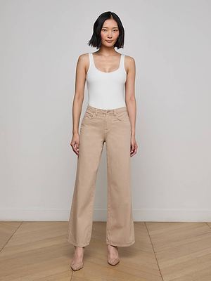 A woman with short black hair models the L'AGENCE Sale Alicent Wide-Leg Jean in beige, styled with a white sleeveless top and beige heels, standing against a light gray wall on a wooden floor.