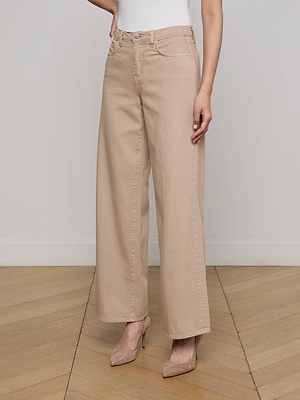 A person wears the L'AGENCE Sale Alicent Wide-Leg Jean in beige with pointed-toe heels, standing against a light-colored wall and wooden floor. The modern sneaker-length pants offer versatile styling. Upper body is partially out of frame.