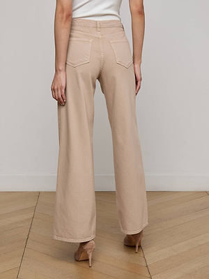 A person wearing the Alicent Wide-Leg Jean by L'AGENCE Sale with a tucked-in white top and beige high heels stands on a wooden floor, facing away from the camera. The high-rise jeans feature back pockets.