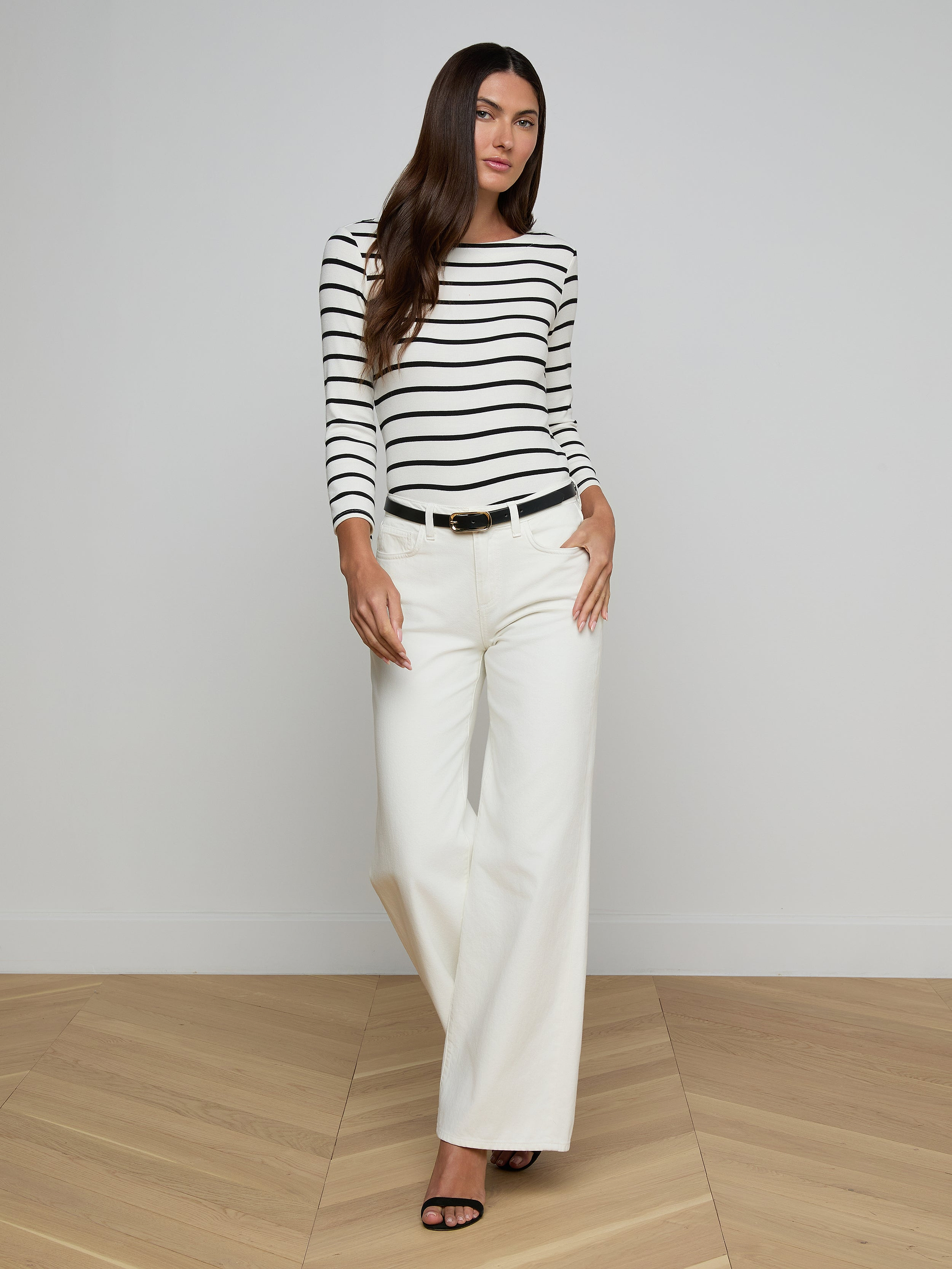A woman stands against a plain background, wearing a black and white striped long-sleeve top, high rise Alicent Wide-Leg Jeans in white, a black belt, and black heels.