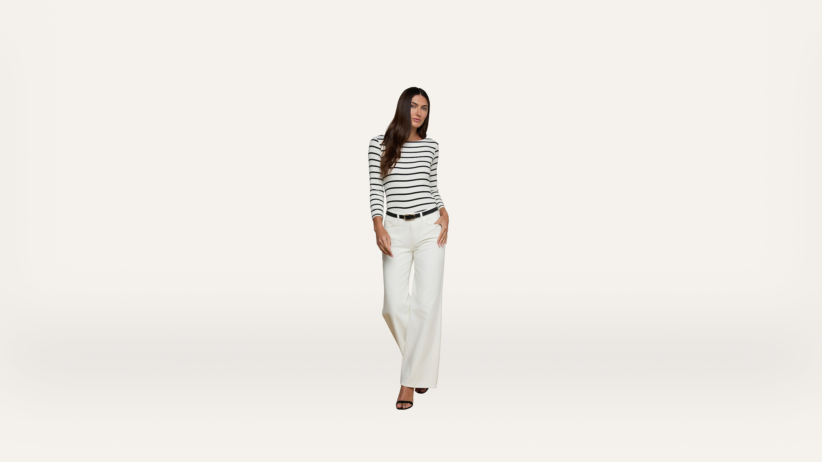 Woman wearing a black-and-white striped long-sleeve shirt, white wide-leg pants, and black heels stands against a plain white background.