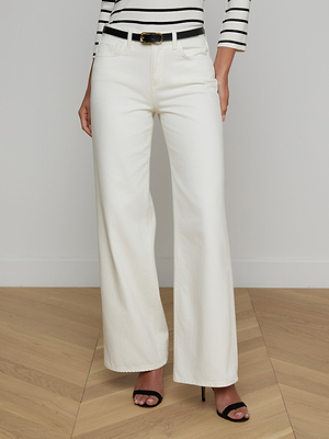 Someone wearing the Alicent Wide-Leg Jean in white, a black belt, a striped long-sleeve top, and black heeled sandals stands on a wooden floor against a plain white wall.