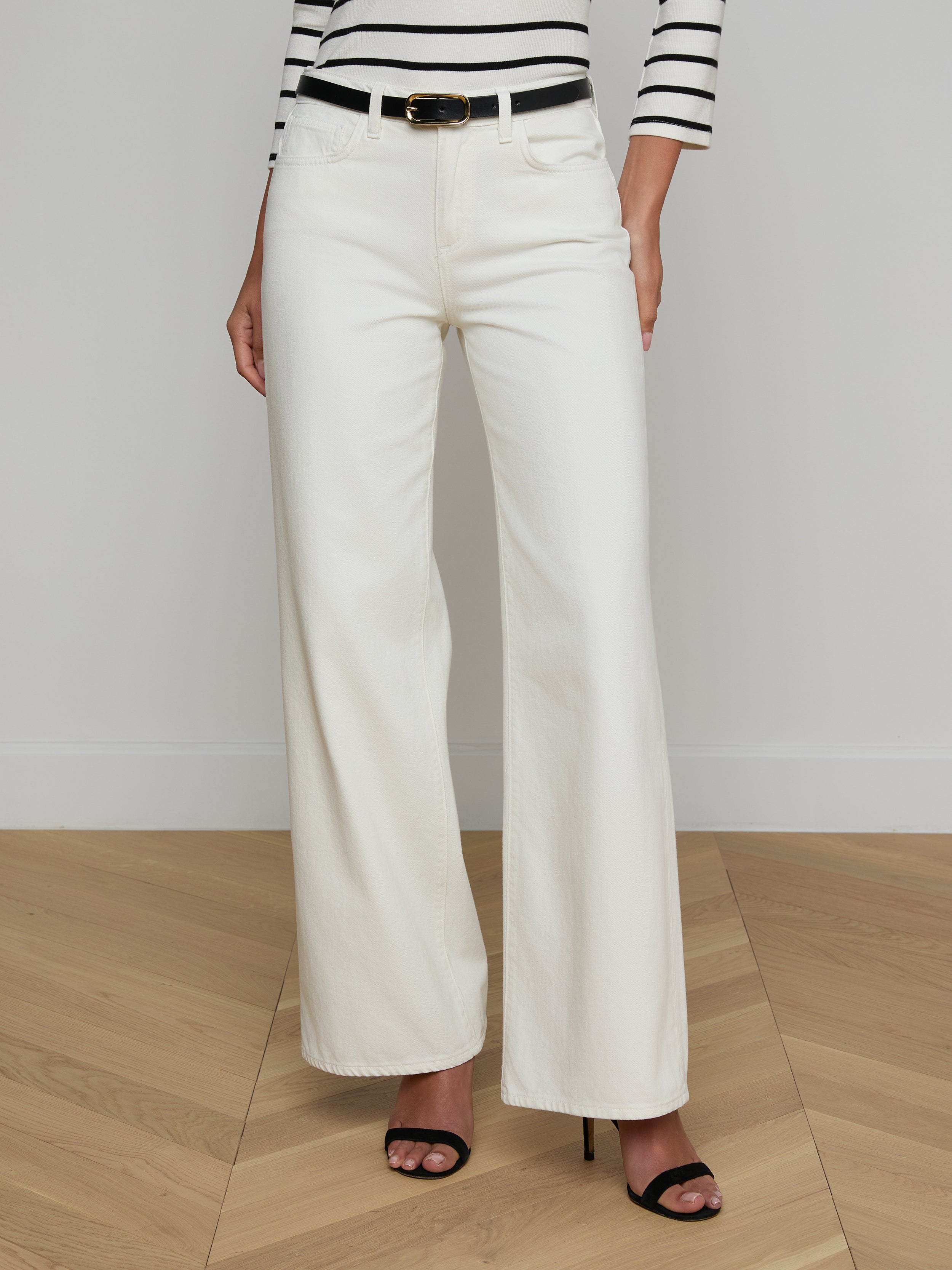 A woman is shown from the shoulders down wearing the Alicent Wide-Leg Jean in white with a high-rise fit, styled with a black belt, black high-heeled sandals, and a black-and-white striped long-sleeve top. The background is plain and light-colored.