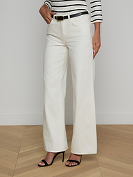 A woman stands against a plain light background wearing the Alicent Wide-Leg Jean in white with a high rise, paired with a black belt, a black and white striped long-sleeve top, and black high-heeled sandals. Only her body is visible; her face is not shown.