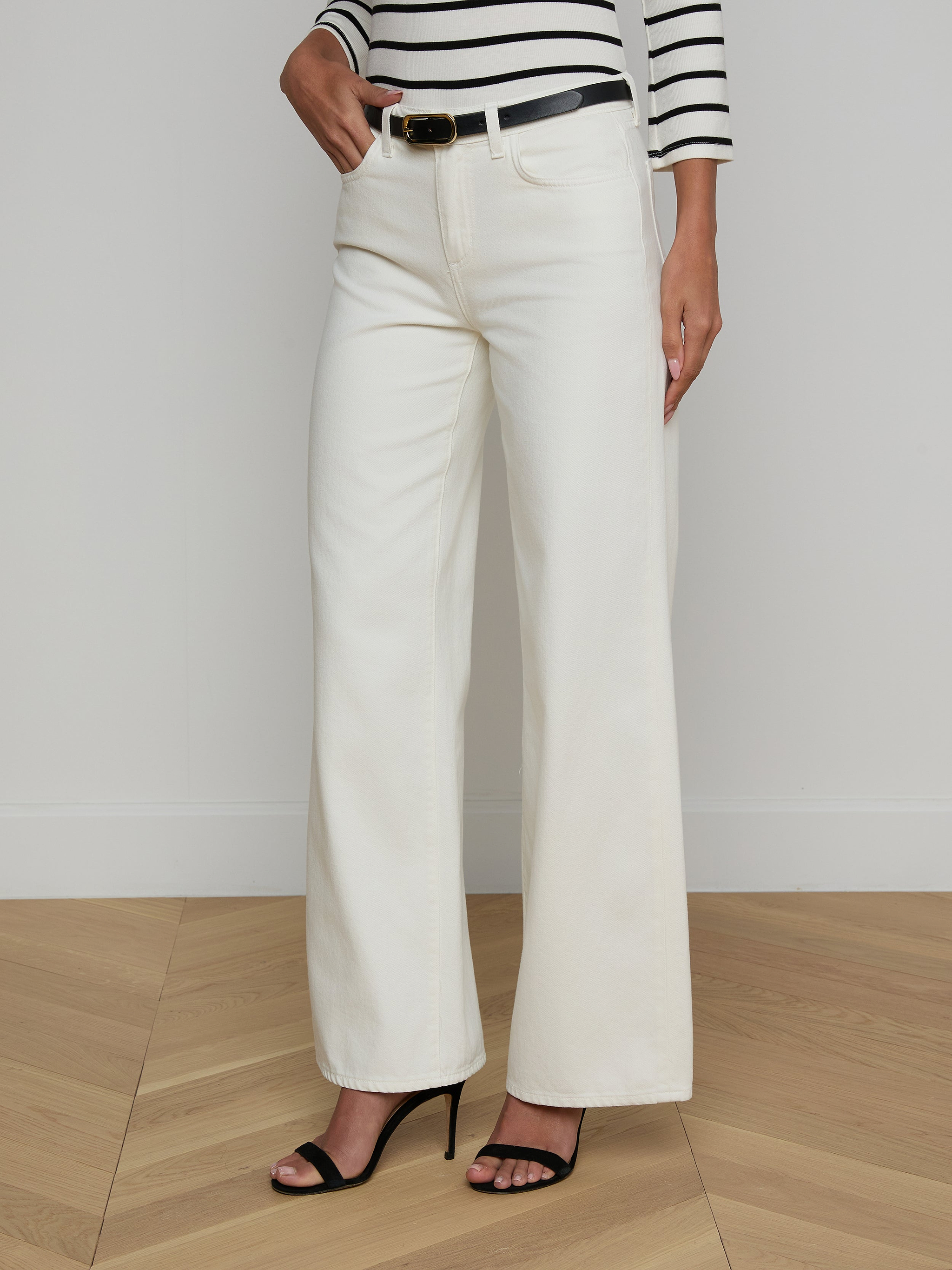A woman stands against a plain light background wearing the Alicent Wide-Leg Jean in white with a high rise, paired with a black belt, a black and white striped long-sleeve top, and black high-heeled sandals. Only her body is visible; her face is not shown.