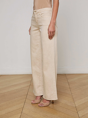 A person wears L'AGENCE Alicent Wide-Leg Jeans in cream subtle stretch denim and beige heels, standing on a light wood floor with a white wall background, shown from shoulders to feet.