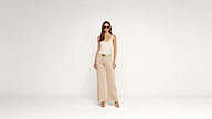 A woman in sunglasses, a white tank top, beige wide-leg pants, and heels stands against a plain white background, facing forward with a relaxed pose.