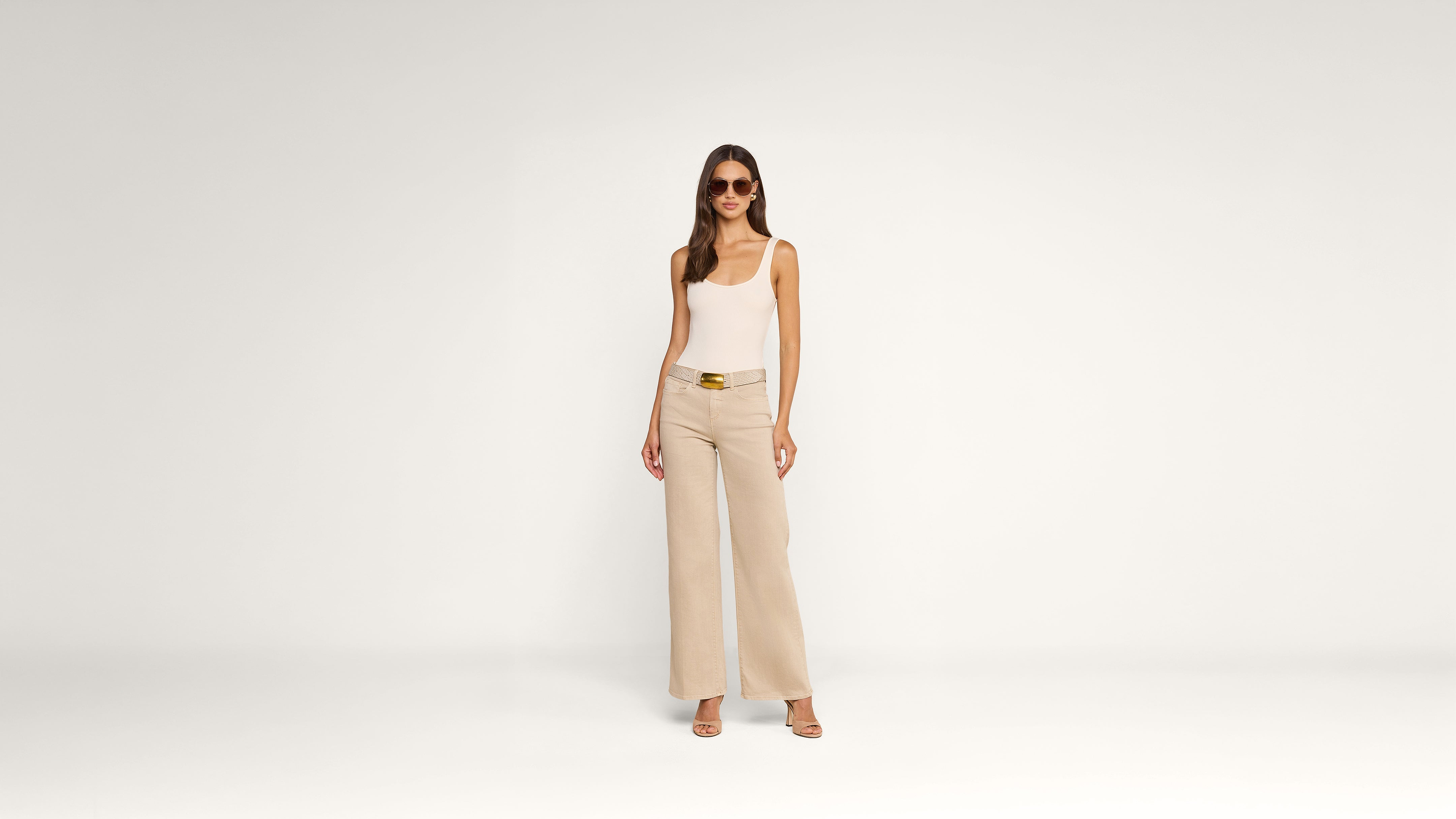 A woman in sunglasses, a white tank top, beige wide-leg pants, and heels stands against a plain white background, facing forward with a relaxed pose.