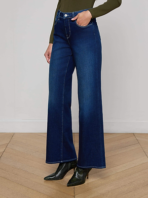Wearing Alicent Wide-Leg Jeans in dark blue stretch denim, a fitted olive green top, and black heeled ankle boots, someone stands on a wooden floor against a plain white wall.