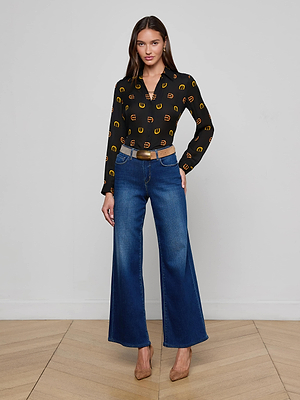 A woman stands indoors on a wooden floor, wearing a black blouse with yellow circles, the L'AGENCE Alicent Wide-Leg Jean in stretch denim, a tan belt, and tan pointed-toe heels against a plain white wall.