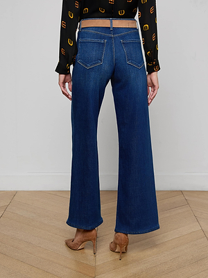 A person stands on a wooden floor facing away, wearing the L'AGENCE Alicent Wide-Leg Jean in stretch blue denim, paired with a black and yellow patterned long-sleeve shirt and brown heeled shoes.