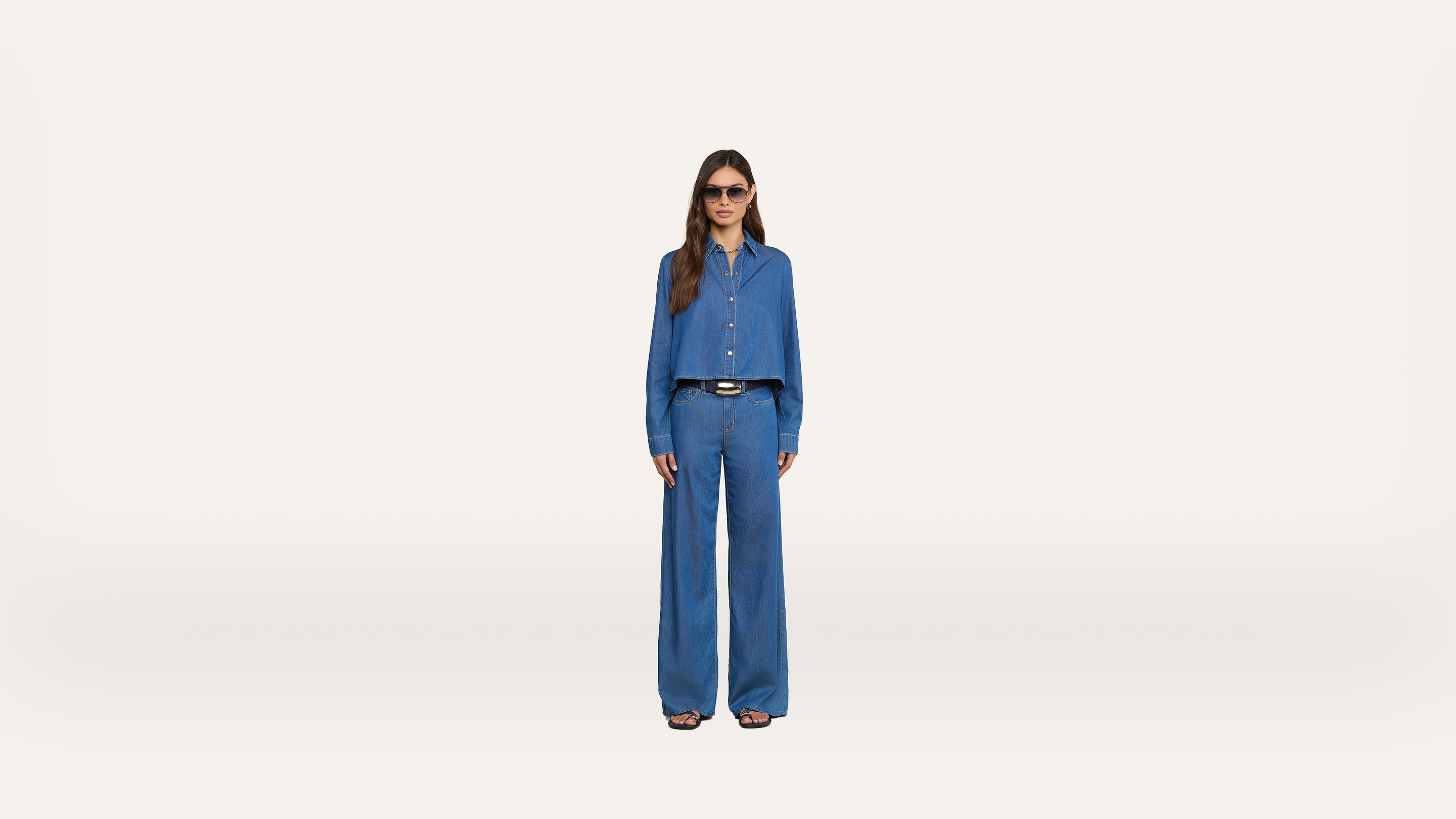 A person wearing sunglasses, a blue button-up shirt, and matching wide-leg blue pants stands facing forward against a plain white background.
