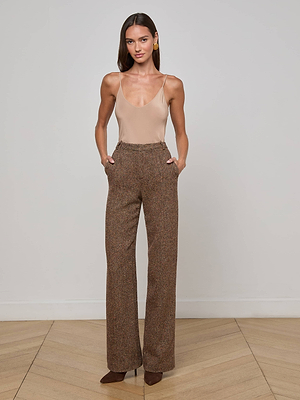 A woman stands against a plain white wall wearing a sleeveless nude bodysuit, L'AGENCE Livvy Straight-Leg Trousers in high-rise Italian recycled wool, and dark brown pointed-toe heels with her hands in her pockets.
