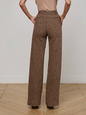 A person stands with hands on hips, wearing L’AGENCE Livvy Straight-Leg Trousers in brown Italian recycled wool and a beige top. The image, taken from behind, highlights the outfit against a neutral wall and wooden floor.