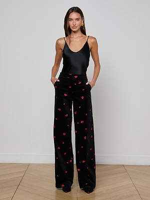 A woman stands against a plain white wall wearing the Livvy Velvet Trouser, a straight-leg black velvet pant embellished with red rose embroidery. She has her hands in her pockets and pairs it with a black sleeveless top.