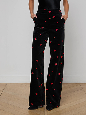 Wearing the Livvy Velvet Trouser—high-waisted, black velvet with red rose embroidery—the model stands with hands in pockets. Wide legs, a tucked-in black top, and pointed shoes complete this chic, elegant look.