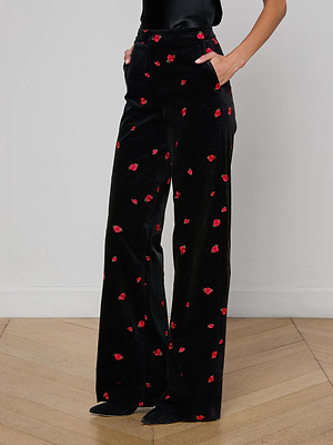 Someone stands on a wooden floor, hands in pockets, wearing the Livvy Velvet Trouser—a high-waisted, straight-leg black velvet pant subtly embroidered with red roses. Only their lower body is visible.