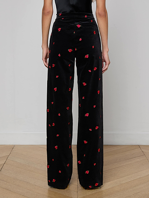 A person stands against a plain wall, shown from the waist down, wearing the Livvy Velvet Trouser—high-waisted black velvet pants with a straight-leg cut and delicate red rose embroidery.