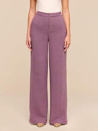 A person stands against a beige background wearing the high-waisted Livvy Straight-Leg Trouser in a purple herringbone pattern paired with beige heels. The top is sleeveless and light-colored, but only the lower half of the body is visible.