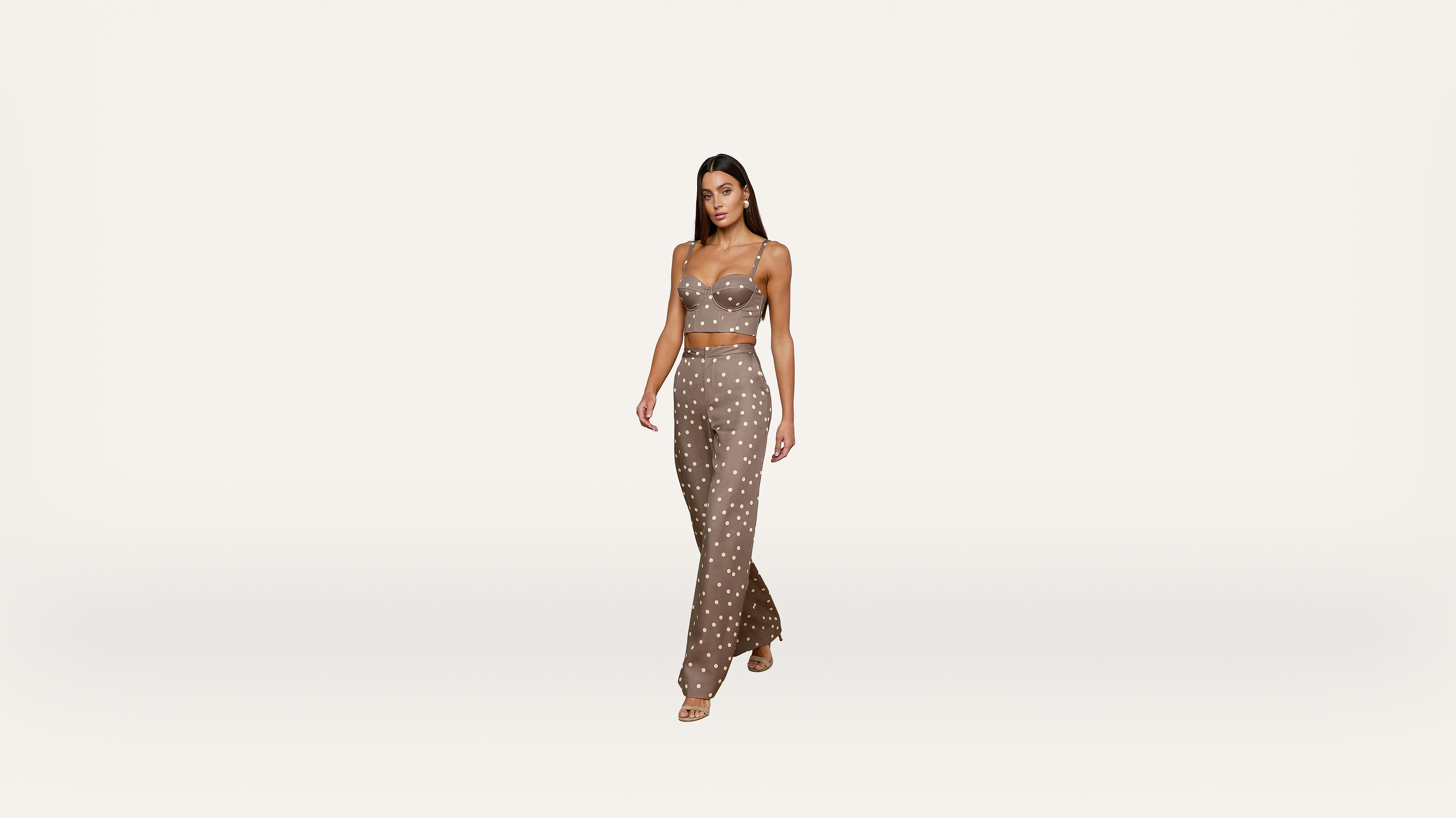 A woman wearing a brown, polka-dot crop top and matching high-waisted pants walks confidently against a plain white background.