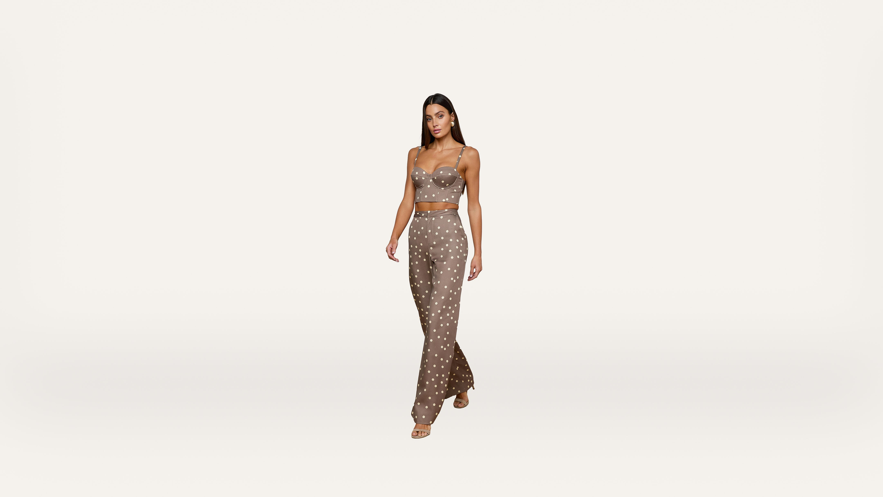 A woman wearing a brown, polka-dot crop top and matching high-waisted pants walks confidently against a plain white background.