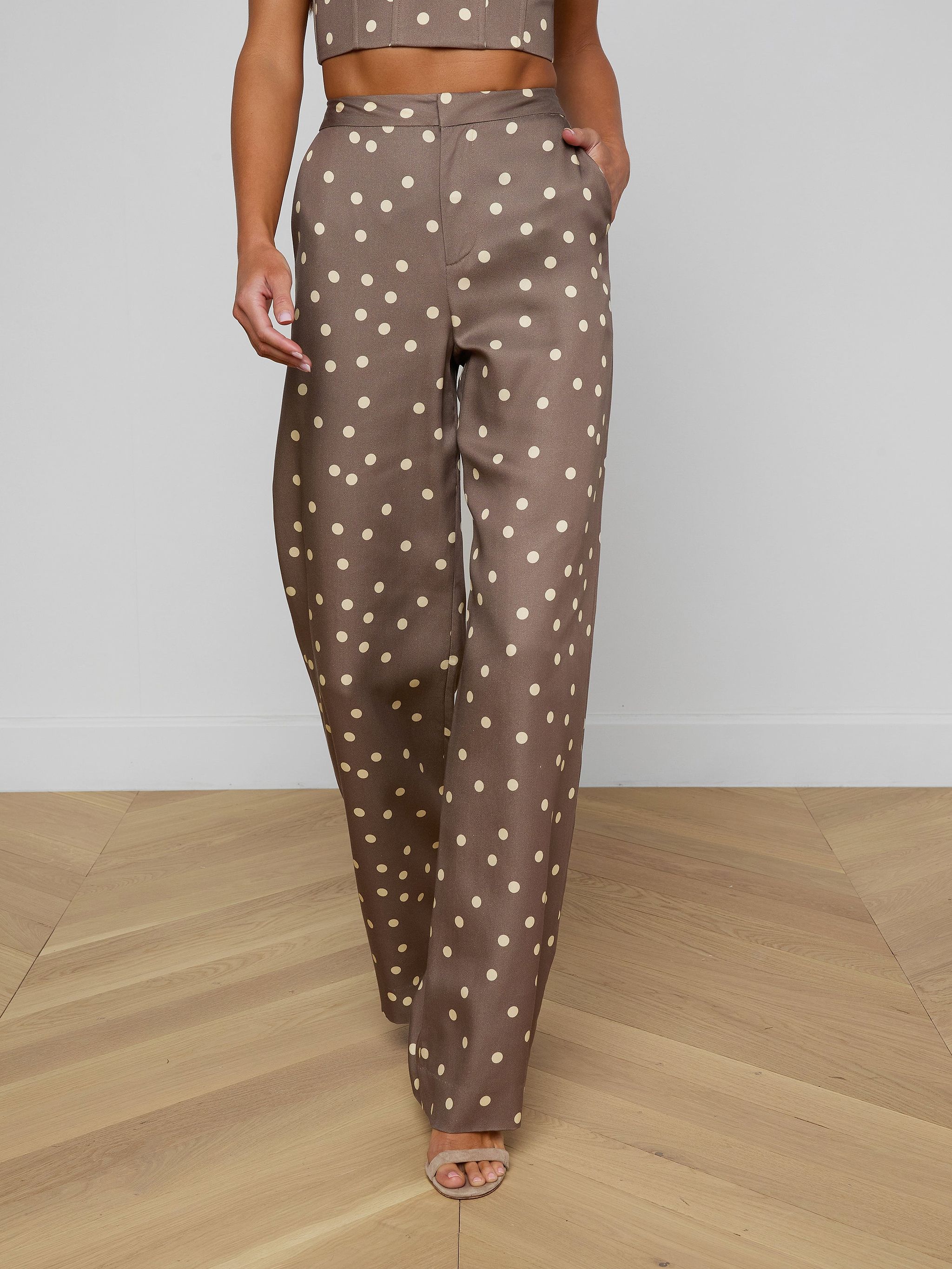 Wearing the Livvy Straight-Leg Trouser featuring a brown polka dot print, a model walks forward against a plain background, styled with a matching crop top and open-toe heels.