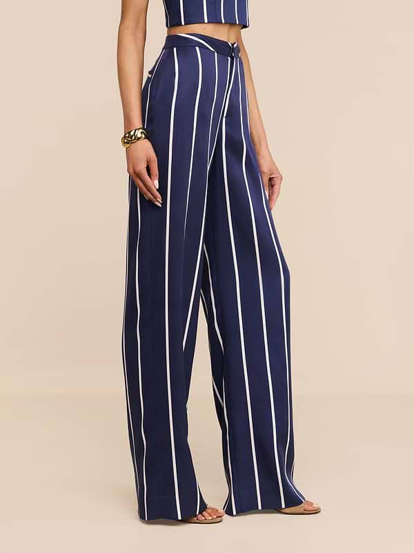 Una persona indossa i Livvy Trouser, pantaloni a vita alta di colore blu con strisce verticali bianche, abbinati a un top coordinato, un braccialetto doro e scarpe aperte, il tutto su uno sfondo beige.