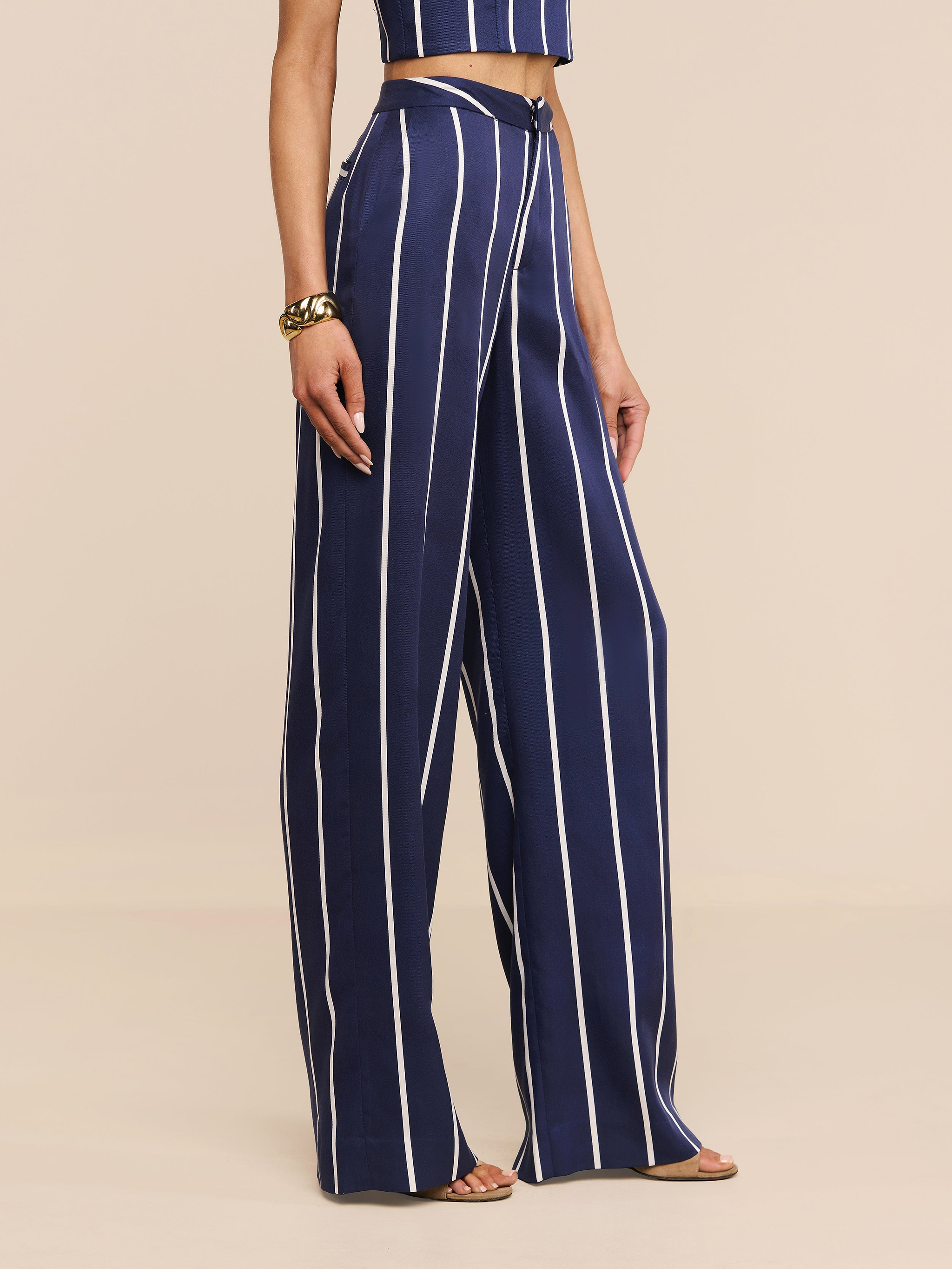 Una persona indossa i Livvy Trouser, pantaloni a vita alta di colore blu con strisce verticali bianche, abbinati a un top coordinato, un braccialetto doro e scarpe aperte, il tutto su uno sfondo beige.