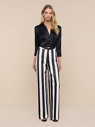 A woman stands against a plain white background, wearing the Livvy Satin Straight-Leg Trouser, which features bold vertical black and white stripes. She pairs it with a black satin blouse, her long blonde hair flowing as she looks directly at the camera.