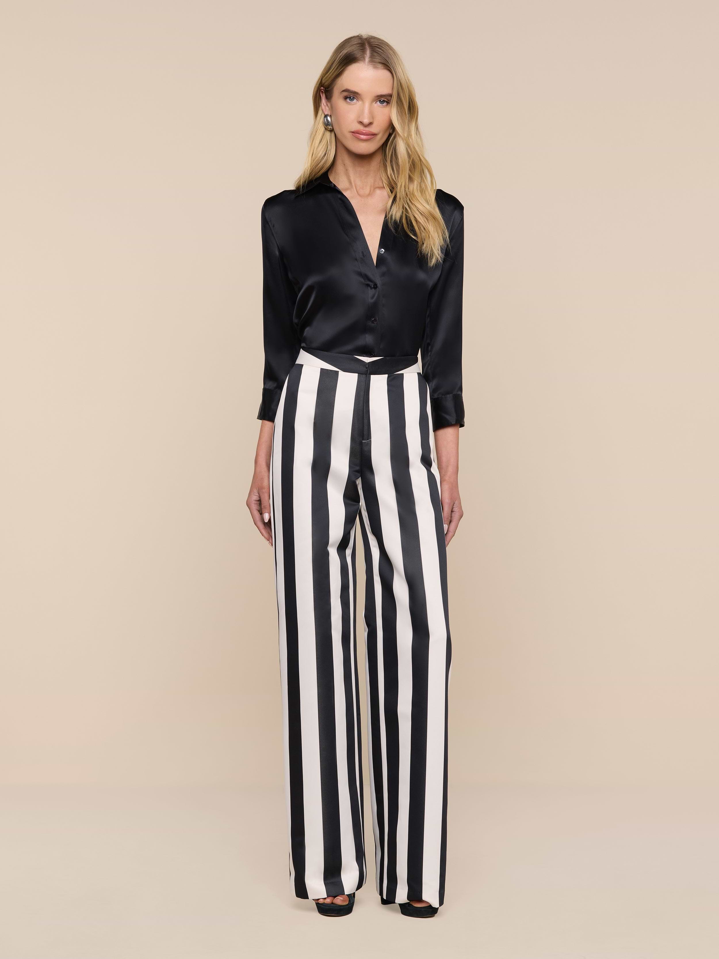 A woman stands against a plain white background, wearing the Livvy Satin Straight-Leg Trouser, which features bold vertical black and white stripes. She pairs it with a black satin blouse, her long blonde hair flowing as she looks directly at the camera.