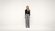 Woman stands against a plain white background wearing a black blouse and high-waisted, wide-leg black and white vertical striped pants. She faces forward with a neutral expression.