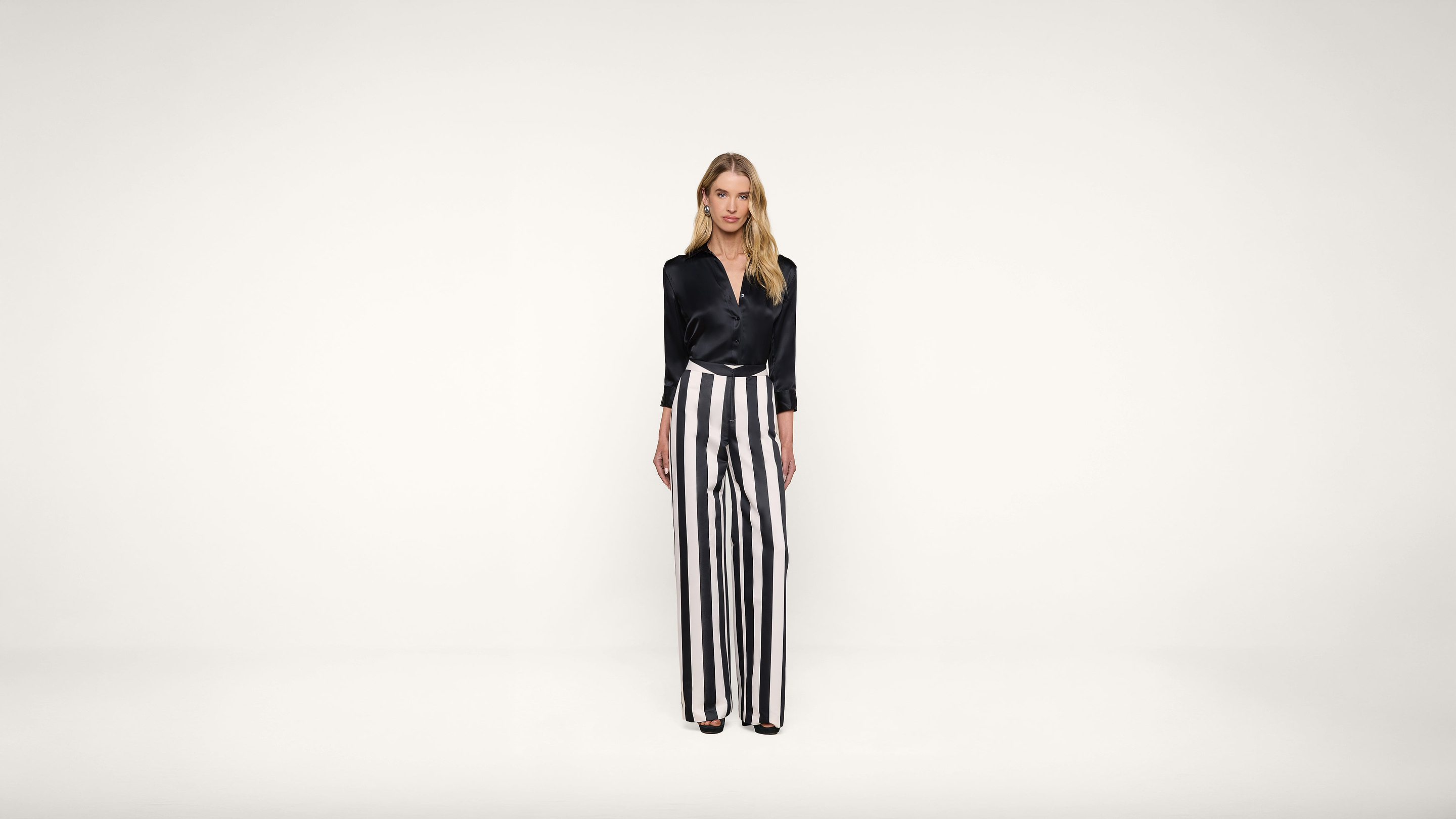 Woman stands against a plain white background wearing a black blouse and high-waisted, wide-leg black and white vertical striped pants. She faces forward with a neutral expression.